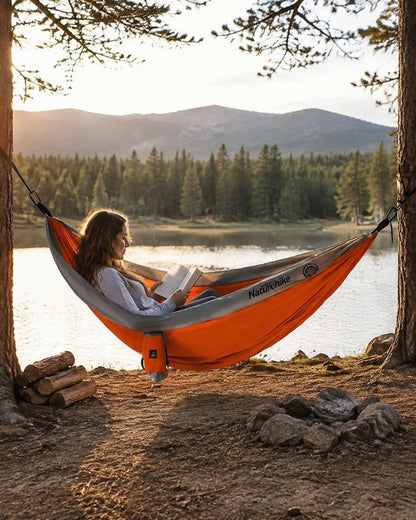 Camper reading in the orange double camping hammock beside a mountain lake