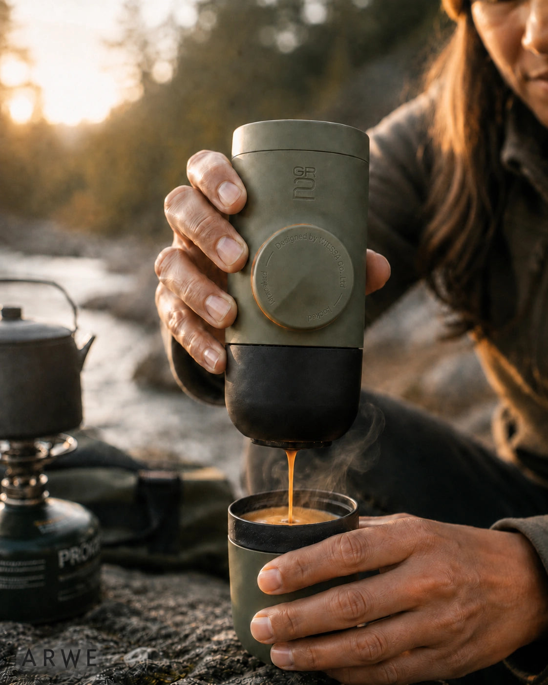Person pouring espresso from a WACACO Minipresso GR2 beside a river while camping